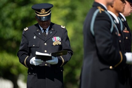 An Army chaplain dressed in dark ceremonial uniform is looking down at a book during a funeral as other soldiers on the right are folding a flag.