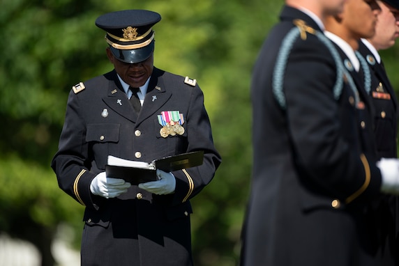 An Army chaplain dressed in dark ceremonial uniform is looking down at a book during a funeral as other soldiers on the right are folding a flag.