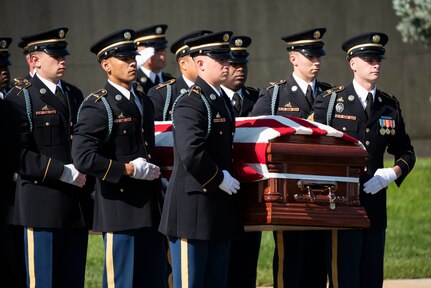 Army Soldiers in dark ceremonial uniforms with white gloves are carrying a casket during a funeral