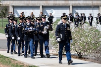 About 10 Army Soldiers dressed in dark ceremonial uniforms are marching in two rows behind another Soldier as they march in a funeral procession at Arlington National Cemetery. There is a columbarium visible in the distance.
