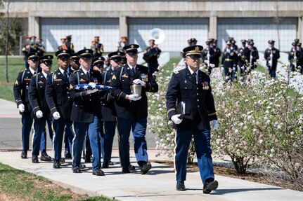 About 10 Army Soldiers dressed in dark ceremonial uniforms are marching in two rows behind another Soldier as they march in a funeral procession at Arlington National Cemetery. There is a columbarium visible in the distance.