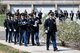 About 10 Army Soldiers dressed in dark ceremonial uniforms are marching in two rows behind another Soldier as they march in a funeral procession at Arlington National Cemetery. There is a columbarium visible in the distance.