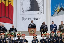 Service members in various types of uniforms are standing in front of and near a podium in a white marble amphitheater. In the background are banners, and one reads "He is risen!"