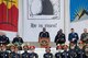 Service members in various types of uniforms are standing in front of and near a podium in a white marble amphitheater. In the background are banners, and one reads "He is risen!"