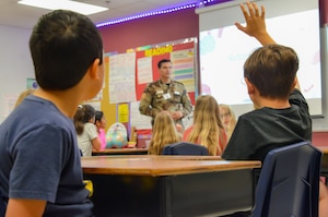 Defense Language Institute English Language Center student talks to a local elementary school class. A student raises their hand to ask a question.