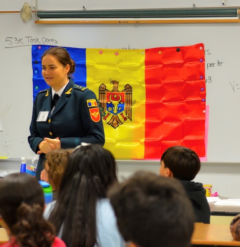Defense Language Institute English Language Center student talks to elementary students. The Moldovan flag is in the background.