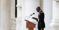 An  Army Chaplain dressed in dark service uniform is holding a book in his hands while looking down at it and reading at a wooden podium in a white-marbled amphitheater.