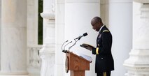 An  Army Chaplain dressed in dark service uniform is holding a book in his hands while looking down at it and reading at a wooden podium in a white-marbled amphitheater.