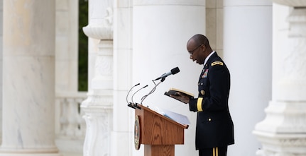 An  Army Chaplain dressed in dark service uniform is holding a book in his hands while looking down at it and reading at a wooden podium in a white-marbled amphitheater.
