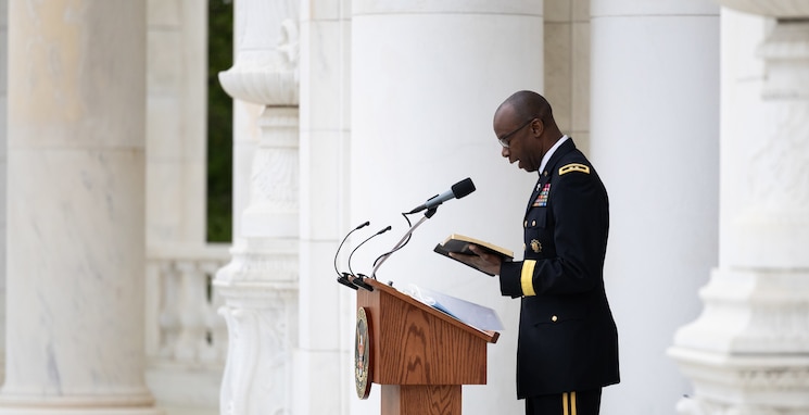 An  Army Chaplain dressed in dark service uniform is holding a book in his hands while looking down at it and reading at a wooden podium in a white-marbled amphitheater.