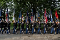 Army Soldiers in dark ceremonial uniforms are marching in a row while carrying flags to form a cordon.
