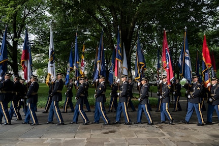 Army Soldiers in dark ceremonial uniforms are marching in a row while carrying flags to form a cordon.