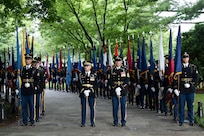Army Soldiers in dark ceremonial uniforms are standing in two rows along a walkway holding various flags in preparation for a flag cordon. Two Soldiers in the middle are holding swords in front of them with the tips pointing into the walkway.