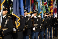 Army Soldiers dressed in dark ceremonial uniforms stand in line for a cordon while carrying various flags.