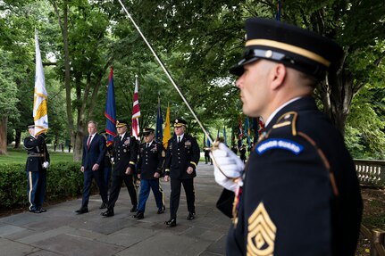 An Army Soldier dressed in ceremonial uniform with an Honor Guard arc on his shoulder above his rank is seen in the foreground holding a sword up in front of his chest as the Secretary of the Army and three Soldiers dressed in Army dress uniforms are walking down a walkway between a flag cordon.