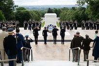 Army Soldiers in various uniforms are facing the white marble Tomb of the Unknown Soldier and saluting. In the middle are four people closer to the tomb, one in civilian attire, who are saluting or placing their hand over their heart in front of a wreath that is placed on a stand.