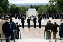 Army Soldiers in various uniforms are facing the white marble Tomb of the Unknown Soldier and saluting. In the middle are four people closer to the tomb, one in civilian attire, who are saluting or placing their hand over their heart in front of a wreath that is placed on a stand.