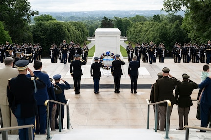 Army Soldiers in various uniforms are facing the white marble Tomb of the Unknown Soldier and saluting. In the middle are four people closer to the tomb, one in civilian attire, who are saluting or placing their hand over their heart in front of a wreath that is placed on a stand.