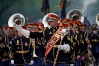 Army Band members dressed in dark uniforms and red hats are marching in formation during a parade while the leader in front is gesturing to the right.