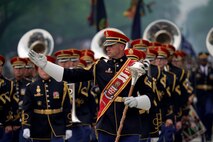 Army Band members dressed in dark uniforms and red hats are marching in formation during a parade while the leader in front is gesturing to the right.