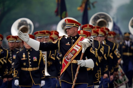 Army Band members dressed in dark uniforms and red hats are marching in formation during a parade while the leader in front is gesturing to the right.
