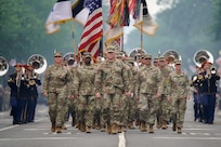 Army Soldiers dressed in combat uniforms are marching in formation down a street during a parade. Some are carrying flags and in the background, member of The U.S. Army Band are performing.