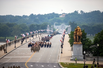 Army Soldiers in various uniforms are seen in the distance marching in serveral formations across a bridge at the start of a parade.