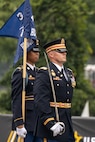 An Army Soldier dressed in dark ceremonial uniform is carrying a sword upright at his side while marching near another Soldier who is carrying a blue flag.