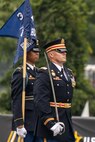 An Army Soldier dressed in dark ceremonial uniform is carrying a sword upright at his side while marching near another Soldier who is carrying a blue flag.