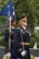 An Army Soldier dressed in dark ceremonial uniform is carrying a sword upright at his side while marching near another Soldier who is carrying a blue flag.