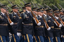 Army Soldiers dressed in dark ceremonial uniforms with white gloves are marching in rows in formation during a parade.