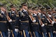 Army Soldiers dressed in dark ceremonial uniforms with white gloves are marching in rows in formation during a parade.