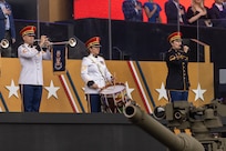An Army Soldier dressed in a white uniform with red hat is playing a herald trumpet next to another Soldier dressed in white uniform who is playing a snare drum. To their left is a Soldier dressed in dark ceremonial uniform holding a microphone and singing. The barrel of a tank is in the foreground.