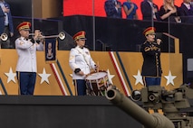 An Army Soldier dressed in a white uniform with red hat is playing a herald trumpet next to another Soldier dressed in white uniform who is playing a snare drum. To their left is a Soldier dressed in dark ceremonial uniform holding a microphone and singing. The barrel of a tank is in the foreground.