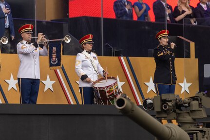 An Army Soldier dressed in a white uniform with red hat is playing a herald trumpet next to another Soldier dressed in white uniform who is playing a snare drum. To their left is a Soldier dressed in dark ceremonial uniform holding a microphone and singing. The barrel of a tank is in the foreground.