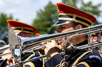 An Army Soldier wearing a red hat and dark ceremonial uniform is playing a trombone while marching in a parade.