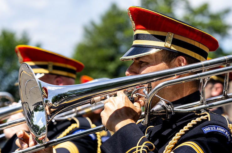 An Army Soldier wearing a red hat and dark ceremonial uniform is playing a trombone while marching in a parade.