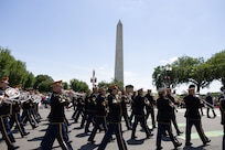 The U.S. Army Band "Pershing's Own" Ceremonial Band is dressed in dark ceremonial uniforms with red hats while marching down a street with the Washington Monument in the background.