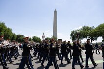 The U.S. Army Band "Pershing's Own" Ceremonial Band is dressed in dark ceremonial uniforms with red hats while marching down a street with the Washington Monument in the background.