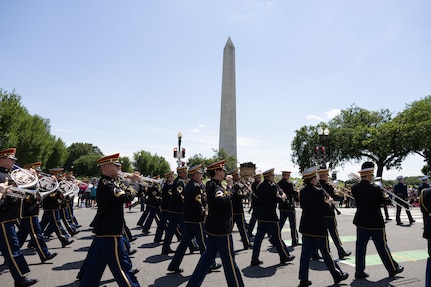The U.S. Army Band "Pershing's Own" Ceremonial Band is dressed in dark ceremonial uniforms with red hats while marching down a street with the Washington Monument in the background.