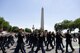 The U.S. Army Band "Pershing's Own" Ceremonial Band is dressed in dark ceremonial uniforms with red hats while marching down a street with the Washington Monument in the background.
