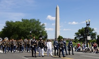 Service members dressed in uniforms from each branch of the military are marching down the street, followed by The U.S. Army Band "Pershing's Own," with the Washington Monument and parade onlookers in the background.
