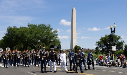 Service members dressed in uniforms from each branch of the military are marching down the street, followed by The U.S. Army Band "Pershing's Own," with the Washington Monument and parade onlookers in the background.