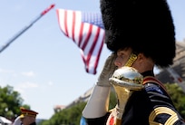Army Band drum major wearing dark ceremonial uniform with large fur-covered hat and carrying a shiny mace in one hand is saluting while marching with a large U.S. flag suspended in the background.