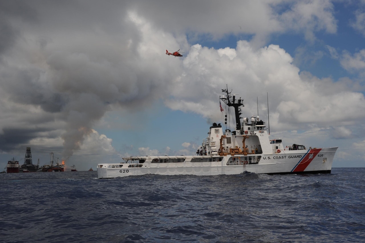 A white ship cruises in the ocean as a helicopter flies overhead near a smoking oil platform.