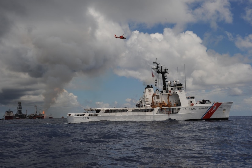 A white ship cruises in the ocean as a helicopter flies overhead near a smoking oil platform.
