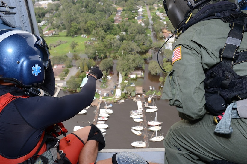 Two people wearing helmets and safety equipment gaze out of an open helicopter door, looking at docked sailing boats near a suburban area.
