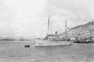 A sailing ship is moored with other ships and a hill is in the background.