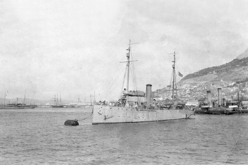 A sailing ship is moored with other ships and a hill is in the background.
