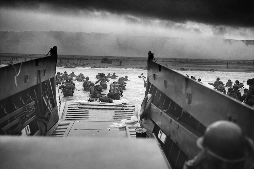 Soldiers in military uniforms wade ashore toward a beach after leaving a landing craft.